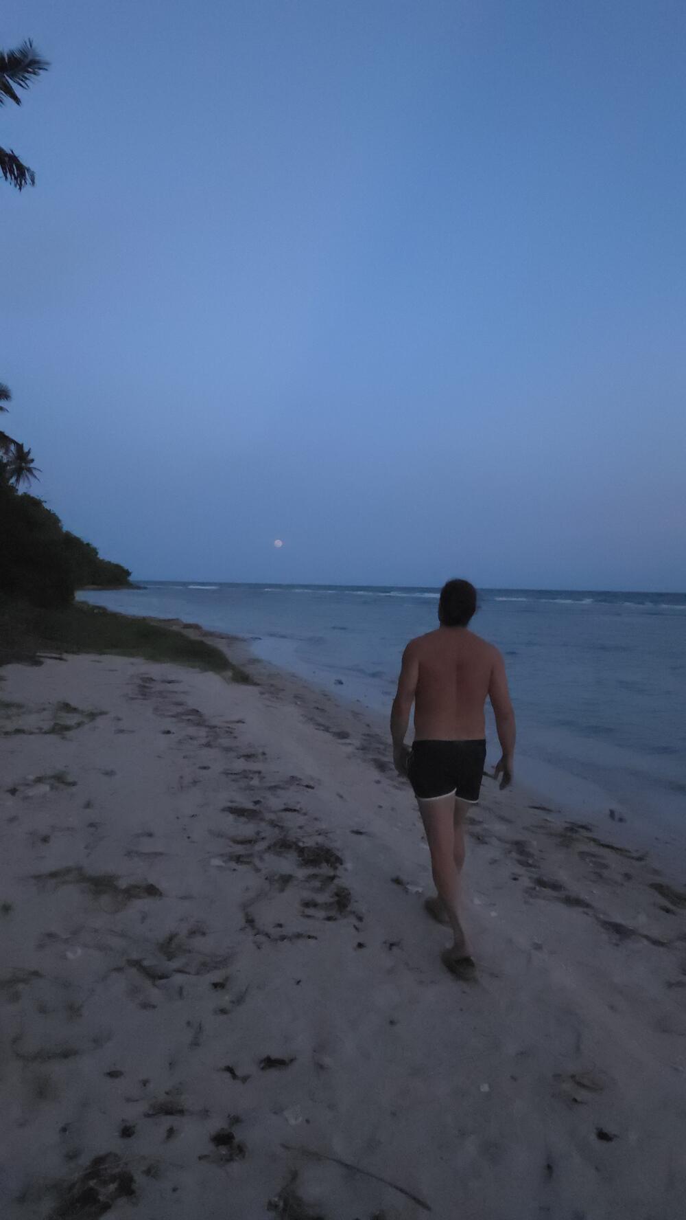 A guest walks barefoot along the shoreline at dusk, waves rolling in.